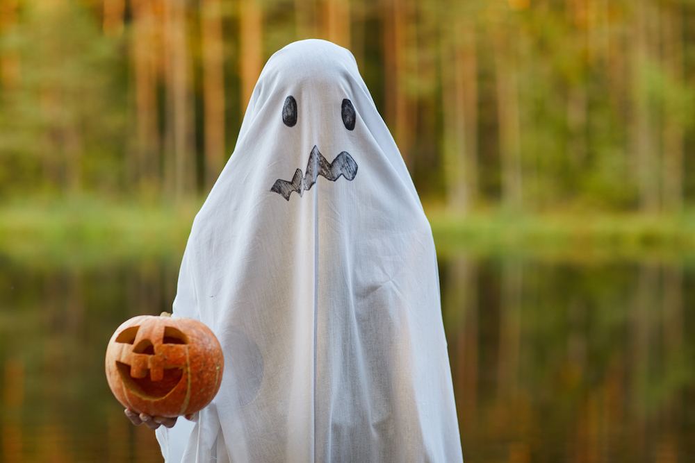 kid dressed as a ghost holding a jack-o-lantern standing by water